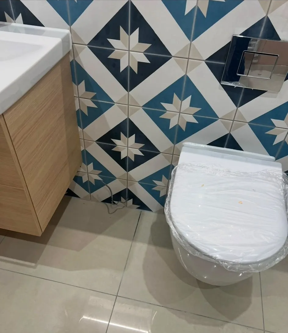 A wall-hung WC against patterned blue and cream tiling, with an oak-effect vanity unit and white basin alongside on polished cream floor tiles.
