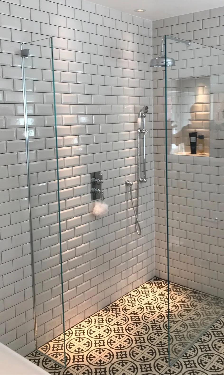 A walk-in shower tiled in white bevelled metro tiles with a chrome rainfall head, a recessed niche and patterned black and white floor tiles.