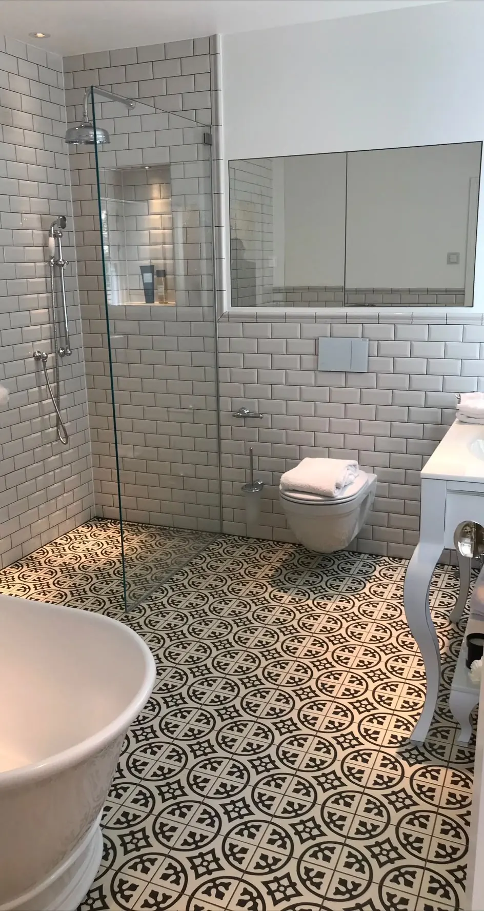 Finished bathroom with a freestanding bath, walk-in shower behind a glass screen, wall-hung WC and white metro tiling over a black and white patterned tiled floor.