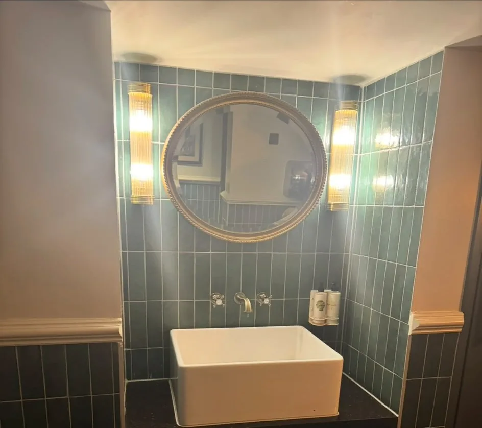 Finished cloakroom with a countertop basin on a dark stone worktop, wall-mounted tap, green vertical tiling and an oval mirror between two ribbed wall lights.