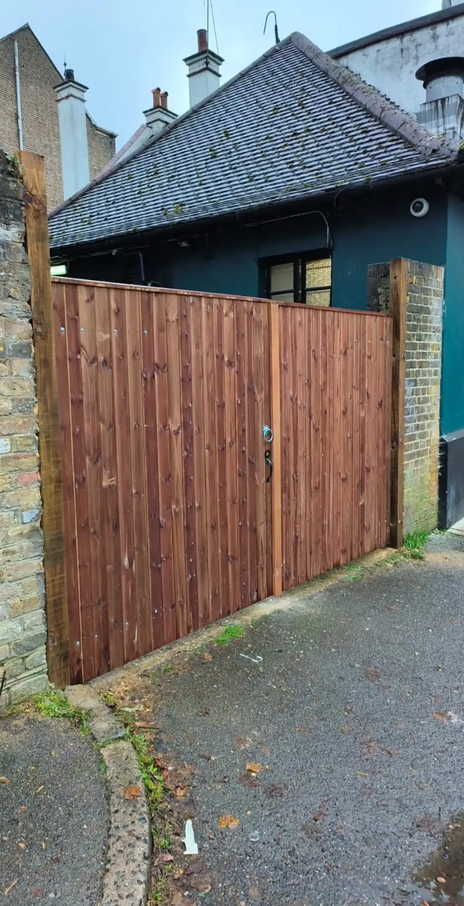 A pair of close-boarded timber gates stained dark brown, hung between brick walls at the entrance to a yard off a back lane.