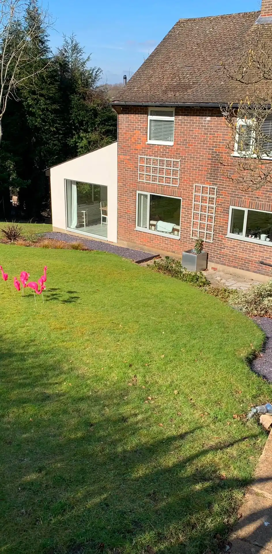 A white rendered single-storey extension with full-width sliding doors on the end of a red brick house, looking back across the lawn.