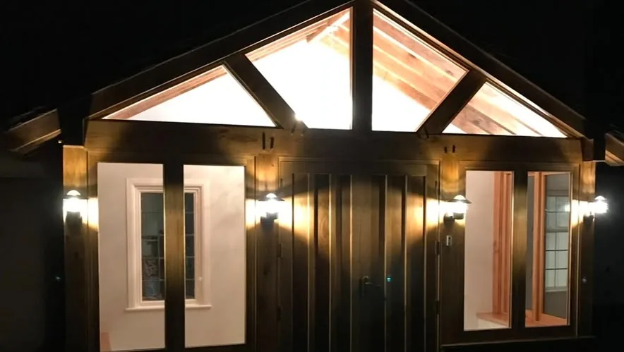 A timber-framed single-storey extension lit at night, with glazed gable above the doors and wall lights along the front.