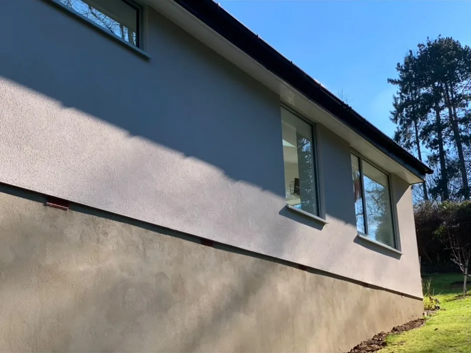 The rendered side elevation of a single-storey house, the lower band of fresh render still darker than the coat above, with turned soil along the foot of the wall.