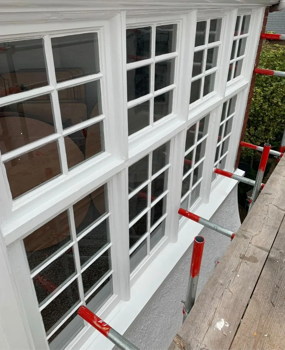 A run of freshly painted white timber windows with glazing bars on a period bay, seen from the scaffold boards alongside.
