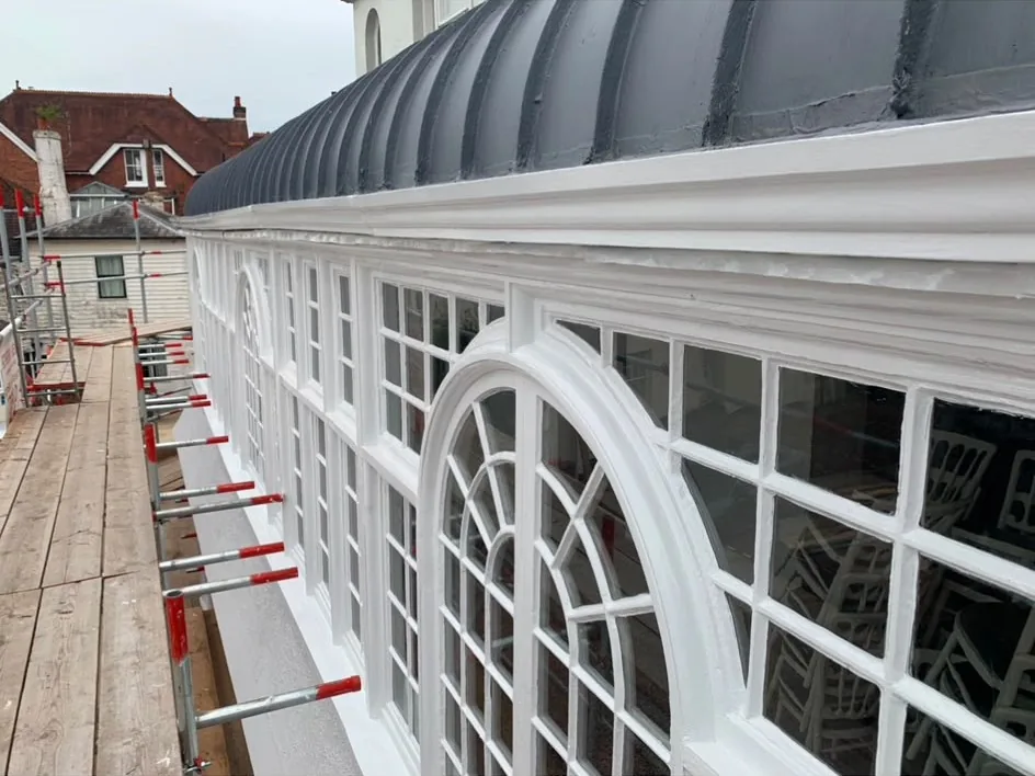 A run of arched multi-pane timber windows on a period building, newly painted white beneath a curved lead roof, seen from the scaffold alongside.