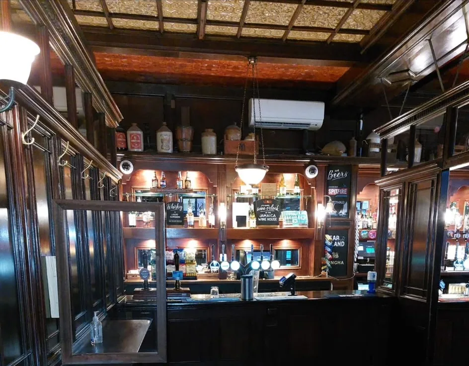 The bar of a traditional pub, with dark mahogany fittings, mirrored spirit shelves, brass hooks on the snug screen and an embossed panel ceiling.
