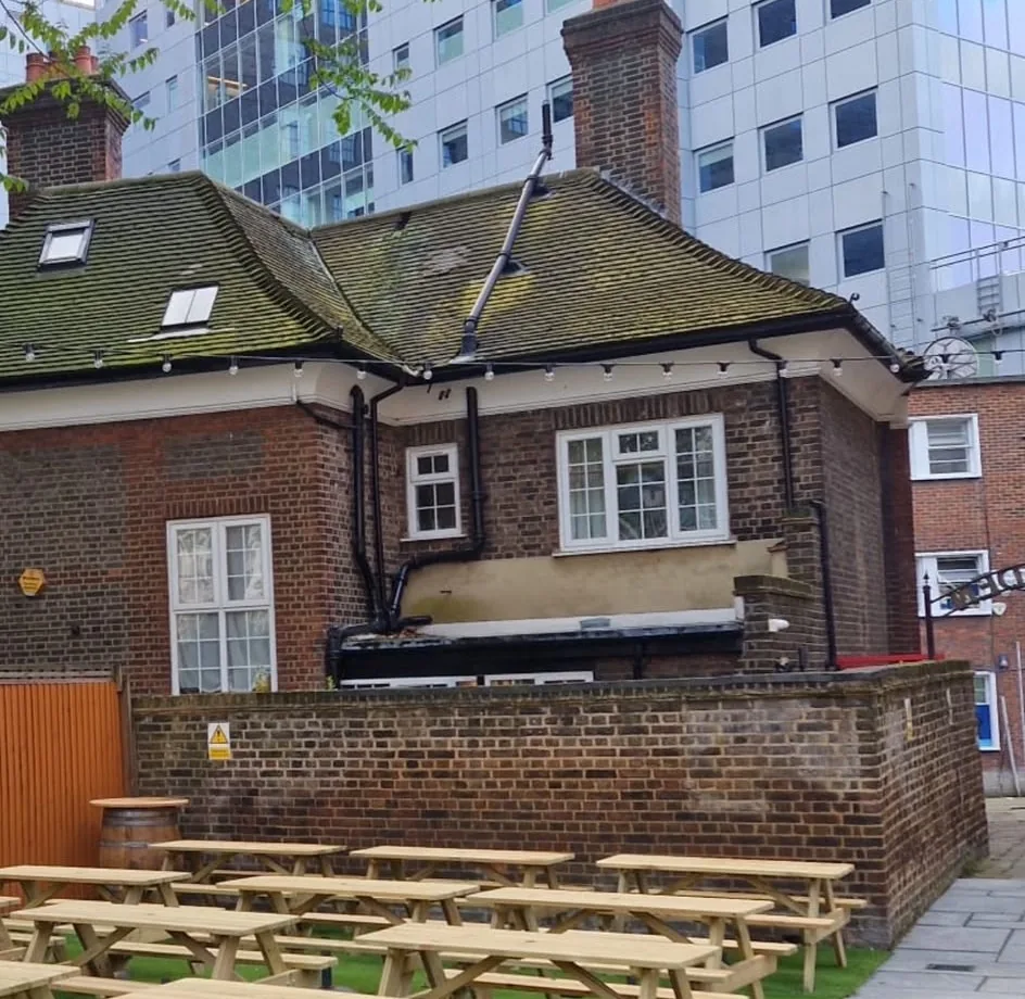 A pub beer garden with new picnic benches on artificial grass, a paved path alongside and festoon lighting run across the brick building behind.