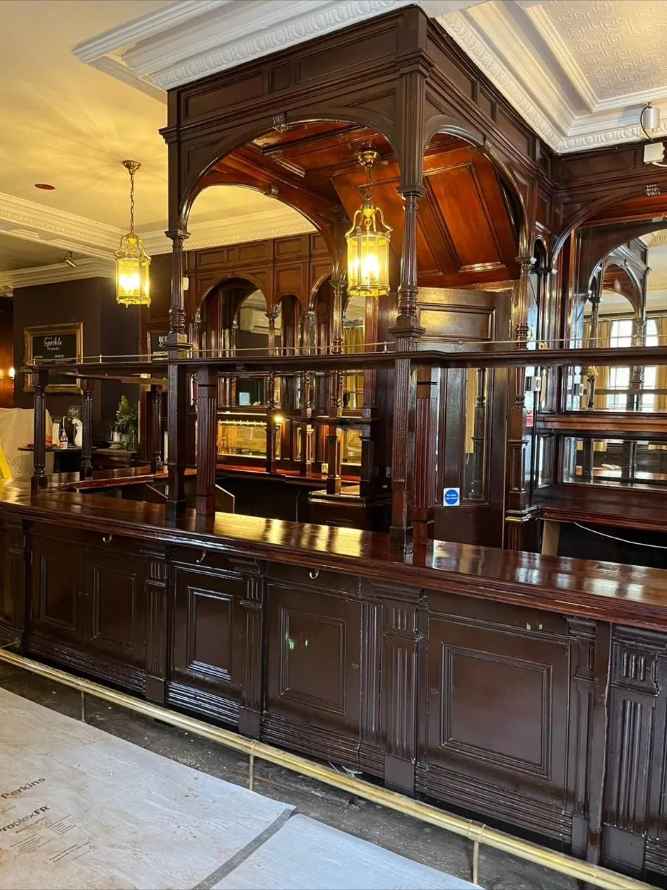 Restored mahogany pub bar with an arched canopy, brass rails and hanging lanterns, with floor protection still down across the boards.