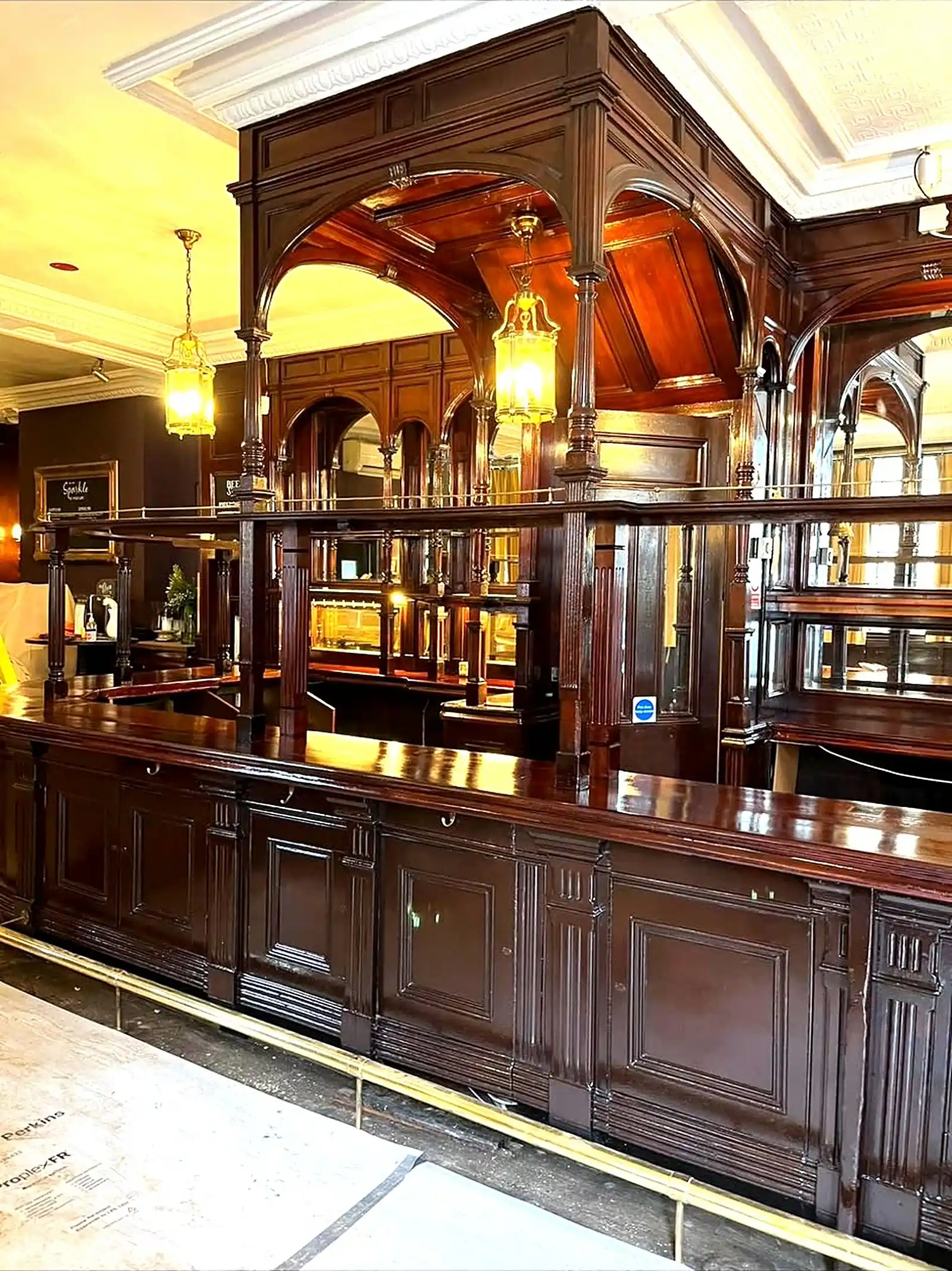 Restored mahogany pub bar with an arched canopy, brass rails and hanging lanterns, with floor protection still down across the boards.