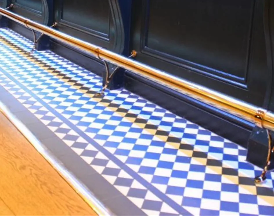 Finished pub bar front in dark painted panelling with a brass foot rail, set on a blue and white chequered tiled floor.