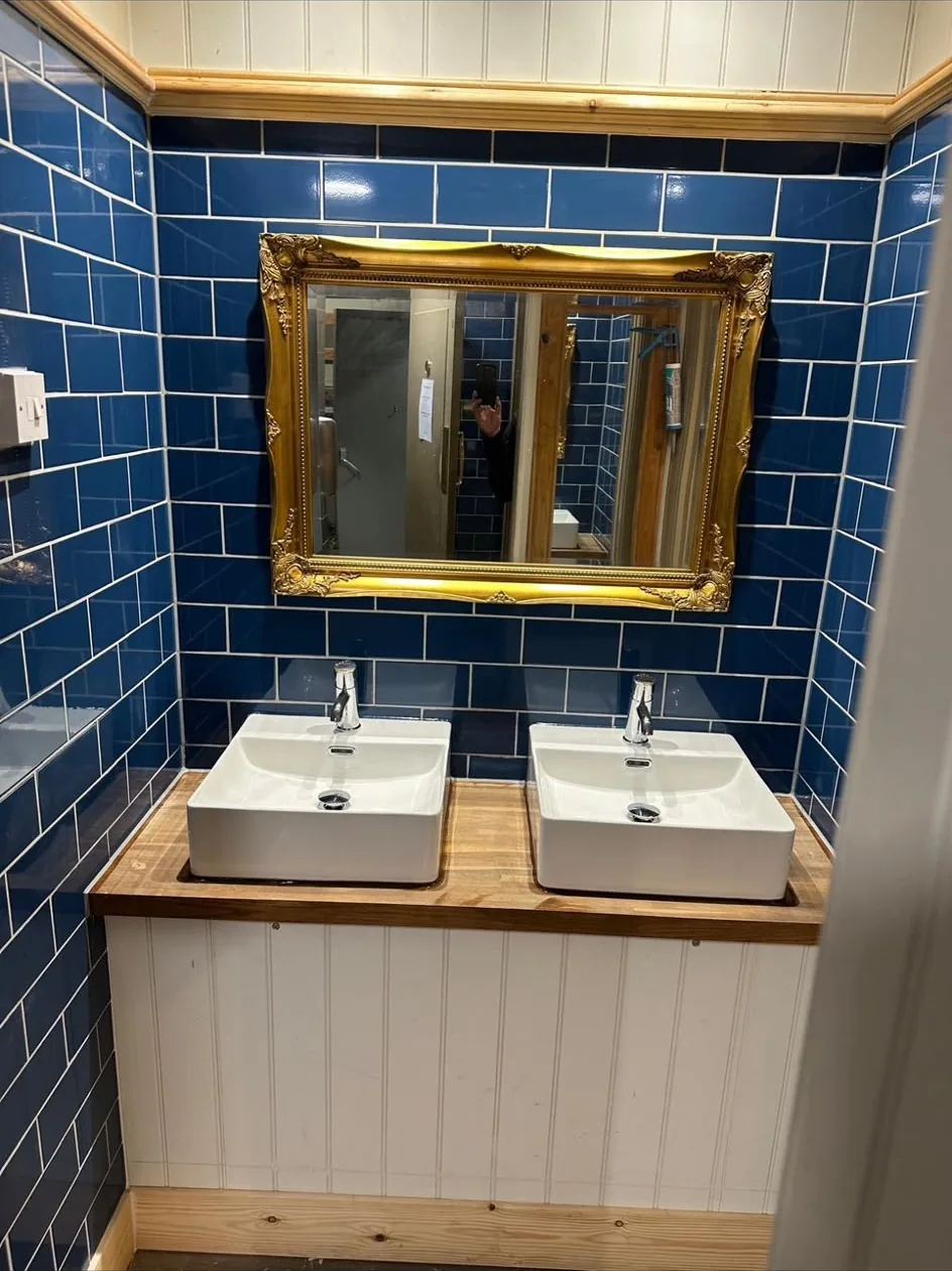Pub washroom with blue metro tiling, twin basins on a timber top over tongue-and-groove panelling, and a gilt mirror.