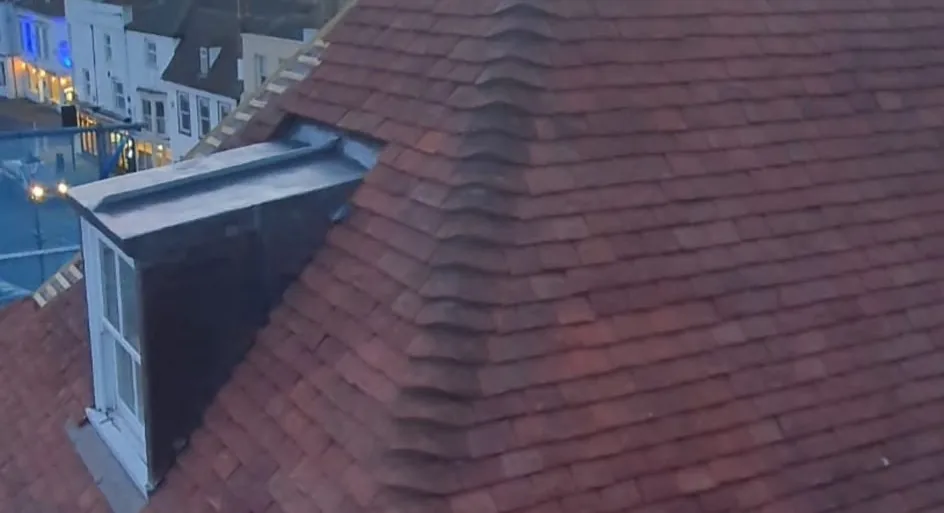 Re-tiled hipped roof in new plain clay tiles with a lead-clad dormer, seen from scaffold level above a town high street at dusk.