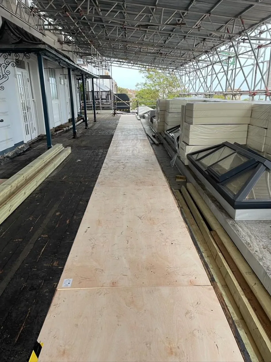 Flat roof stripped back under a temporary scaffold roof, with a plywood walkway laid and insulation boards and new rooflights stacked ready to fit.
