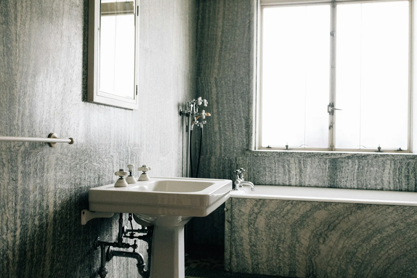 Marbled stone panels reach floor to ceiling behind a pedestal basin with cross-head taps and exposed pipework.