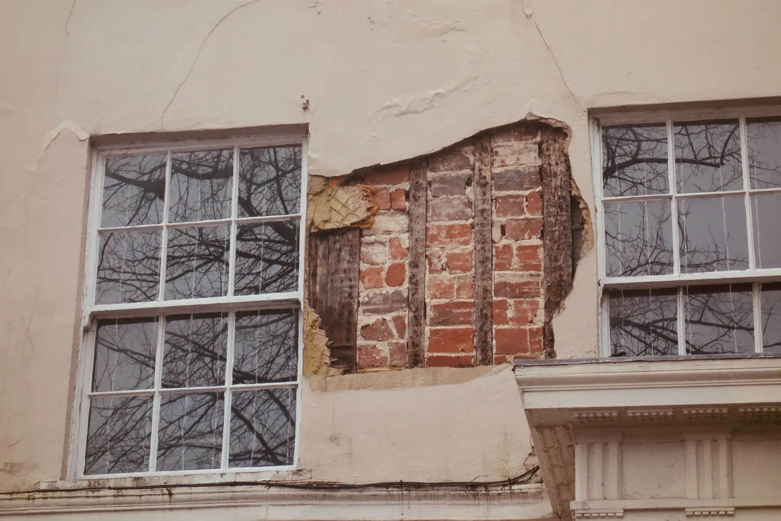 Cracked render fallen away from an old house, exposing timber studwork and brick infill between two sash windows.
