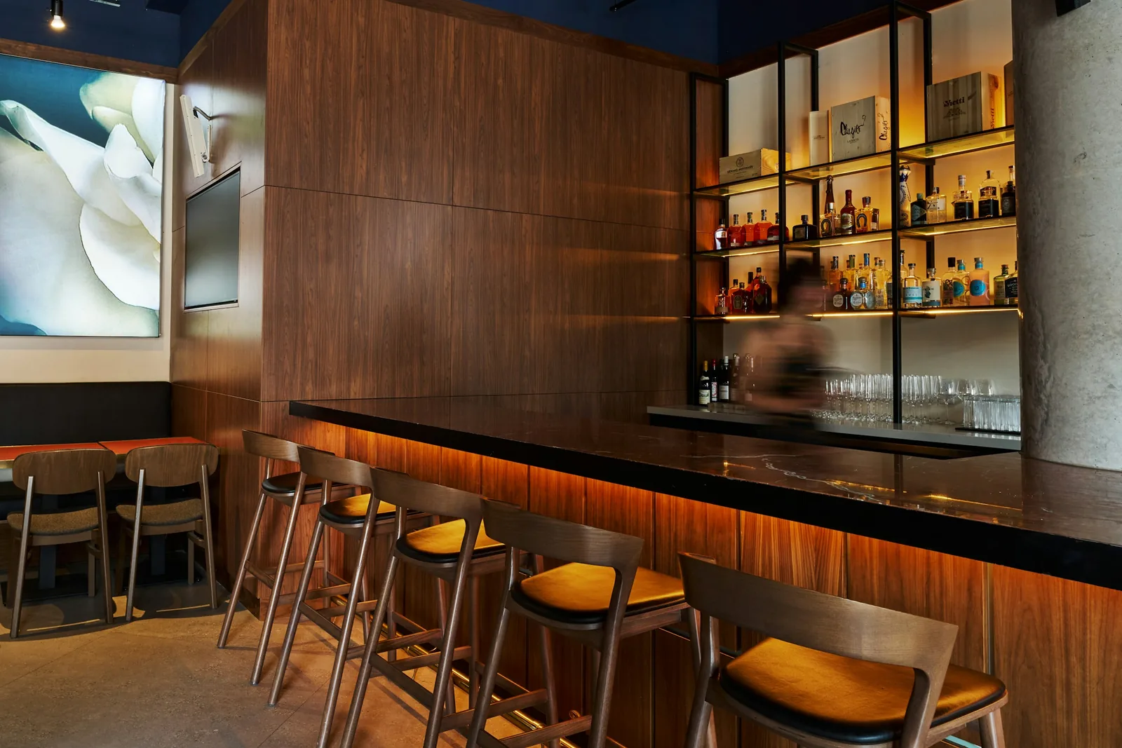 Timber stools line a walnut bar with a dark stone top; the bottle shelving behind it is backlit.