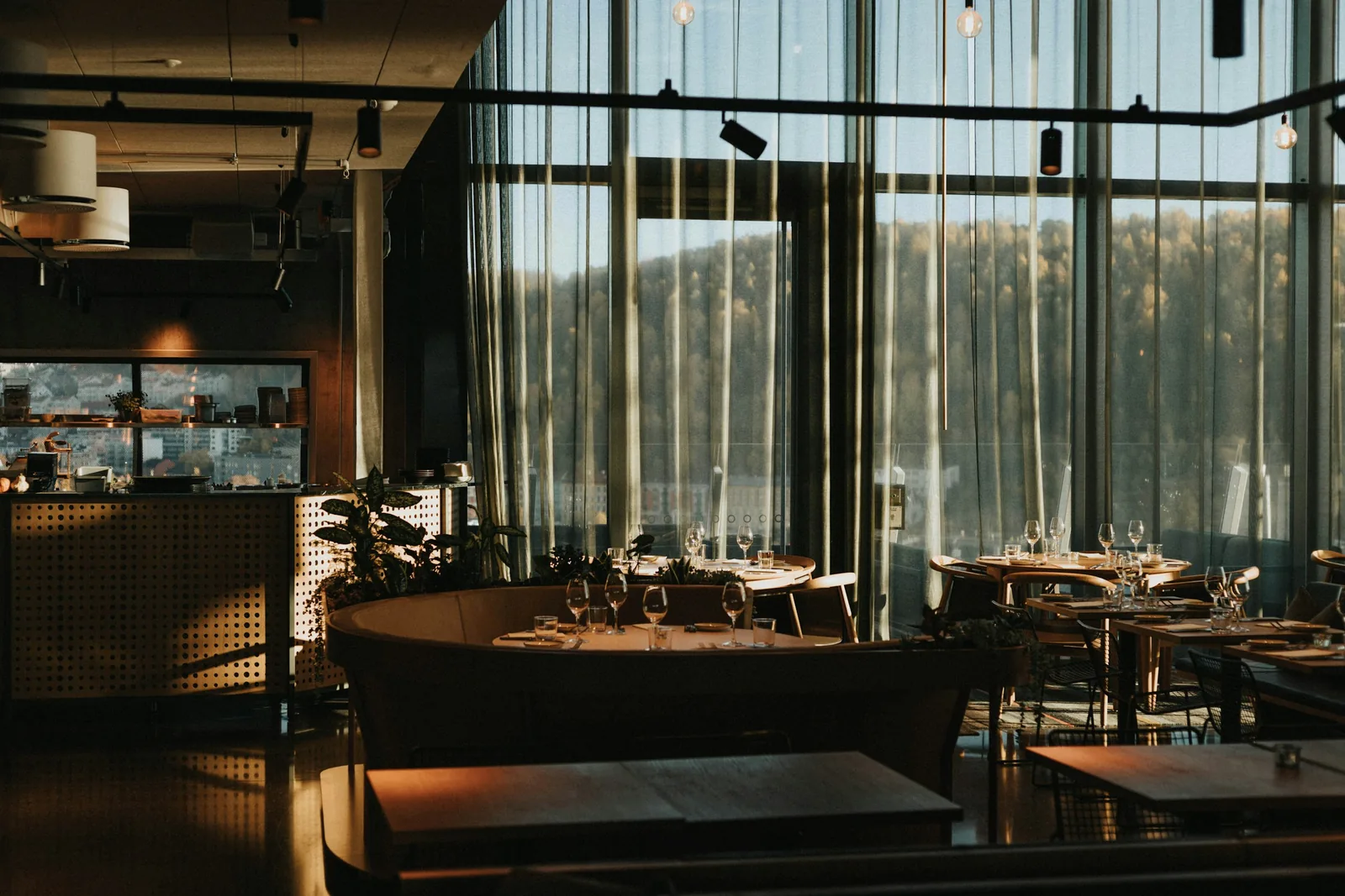 Low sun comes through the sheer curtains onto a curved booth already laid with wine glasses.