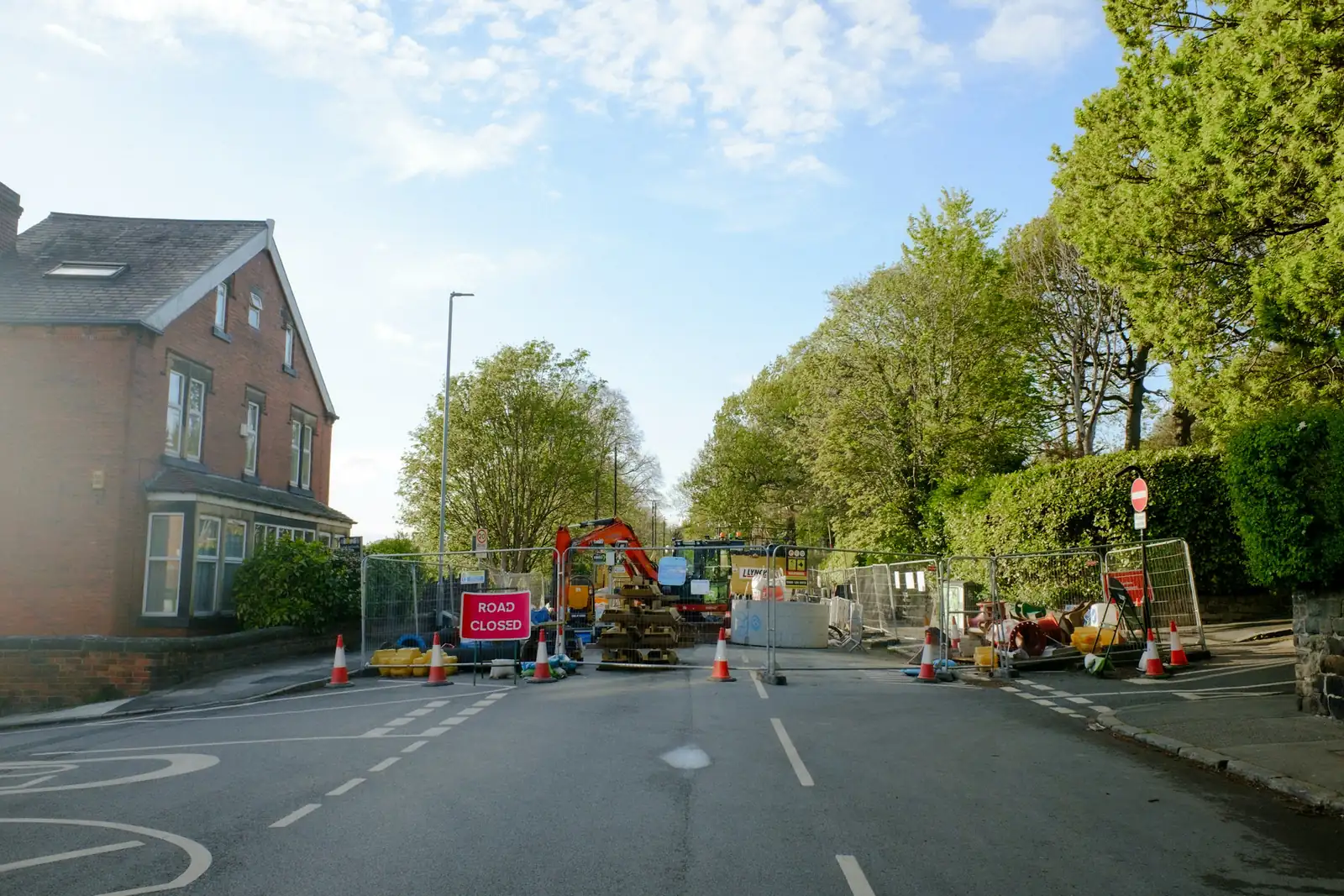 ROAD CLOSED sign and cones across the carriageway, an orange excavator behind the fence panels.