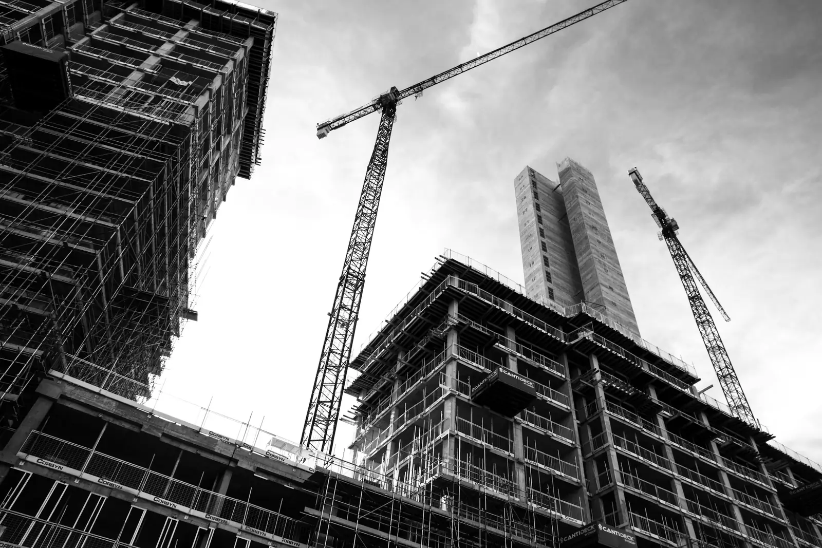 Looking up at two concrete frames in scaffold, with tower cranes and a lift core behind.