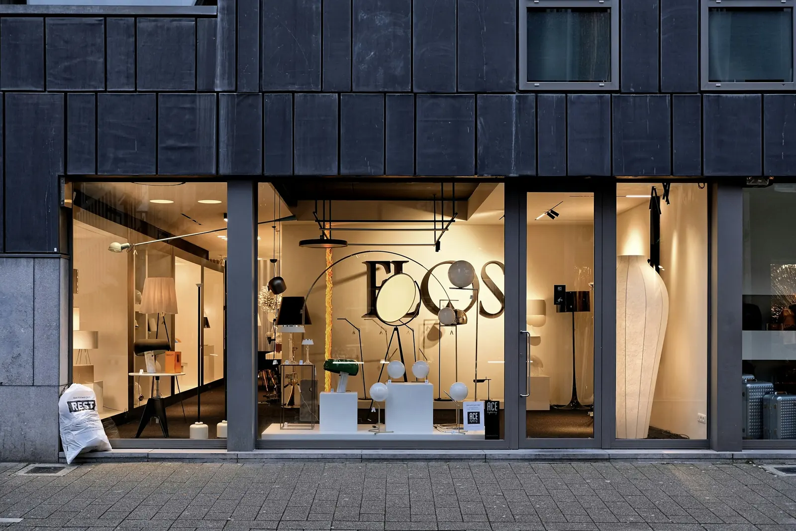 Dark metal cladding above full-height glazing; inside, a lighting showroom lit up at dusk.