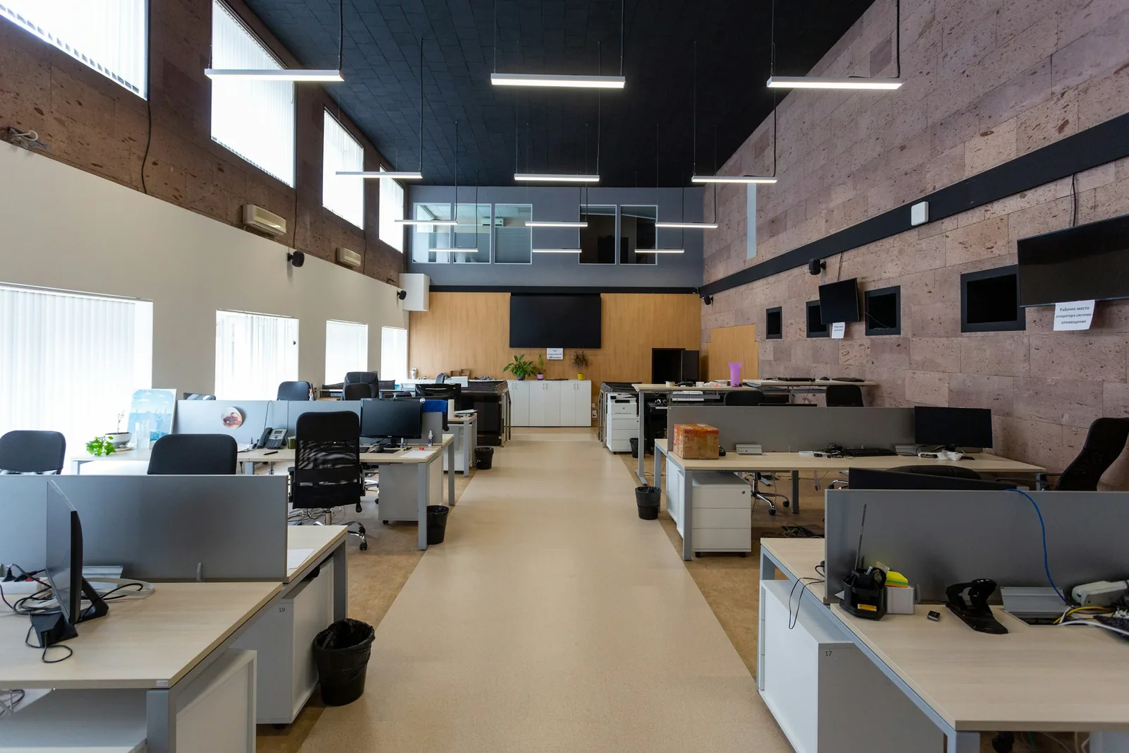 Rows of screened desks under a dark acoustic ceiling, rough stone blockwork on the right wall.