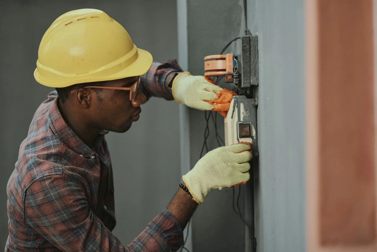 In hard hat and knitted gloves, a fitter works on an electrical box fixed to a column.