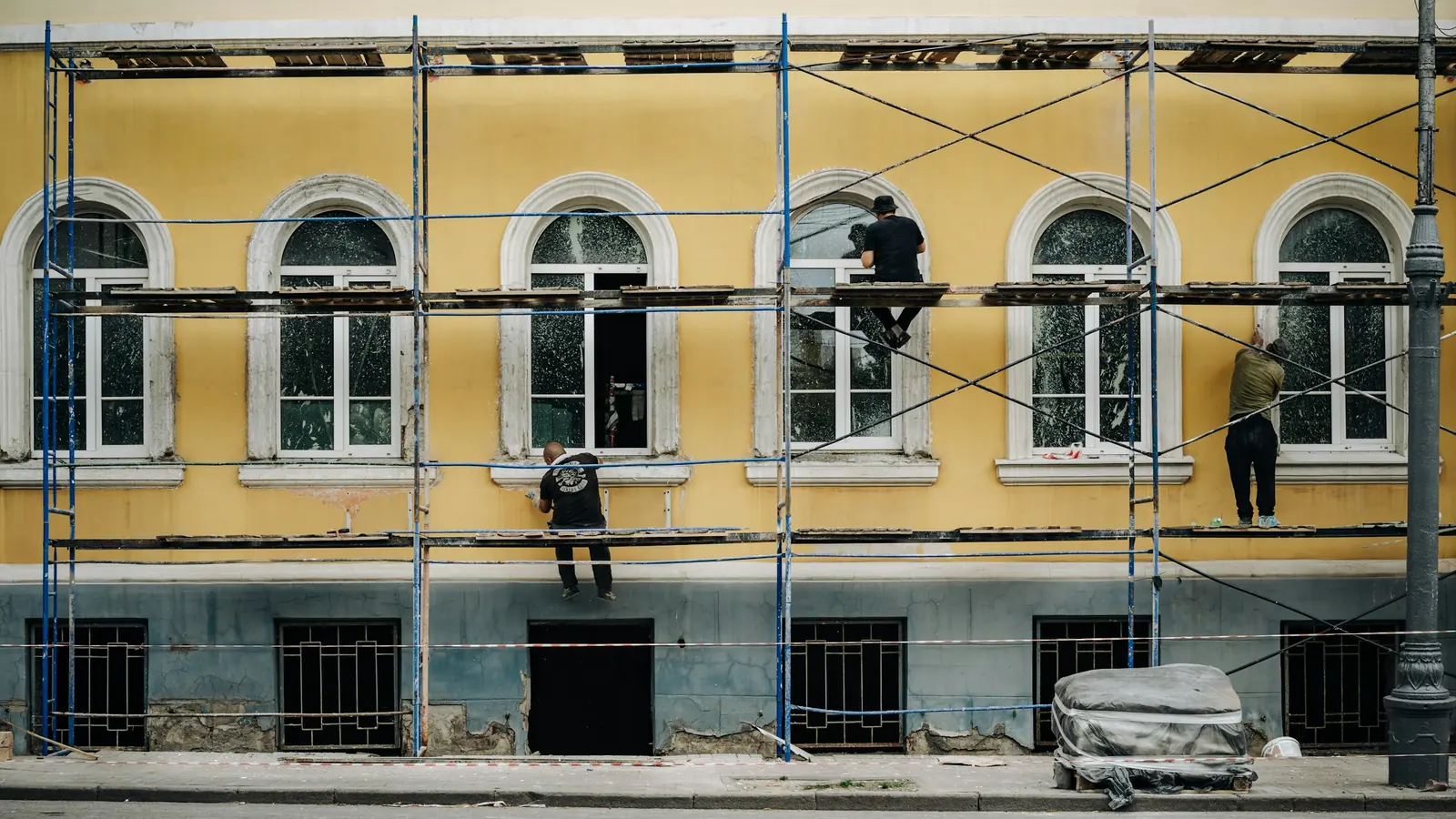 Three men on scaffold boards working across the arched windows of a yellow rendered frontage.