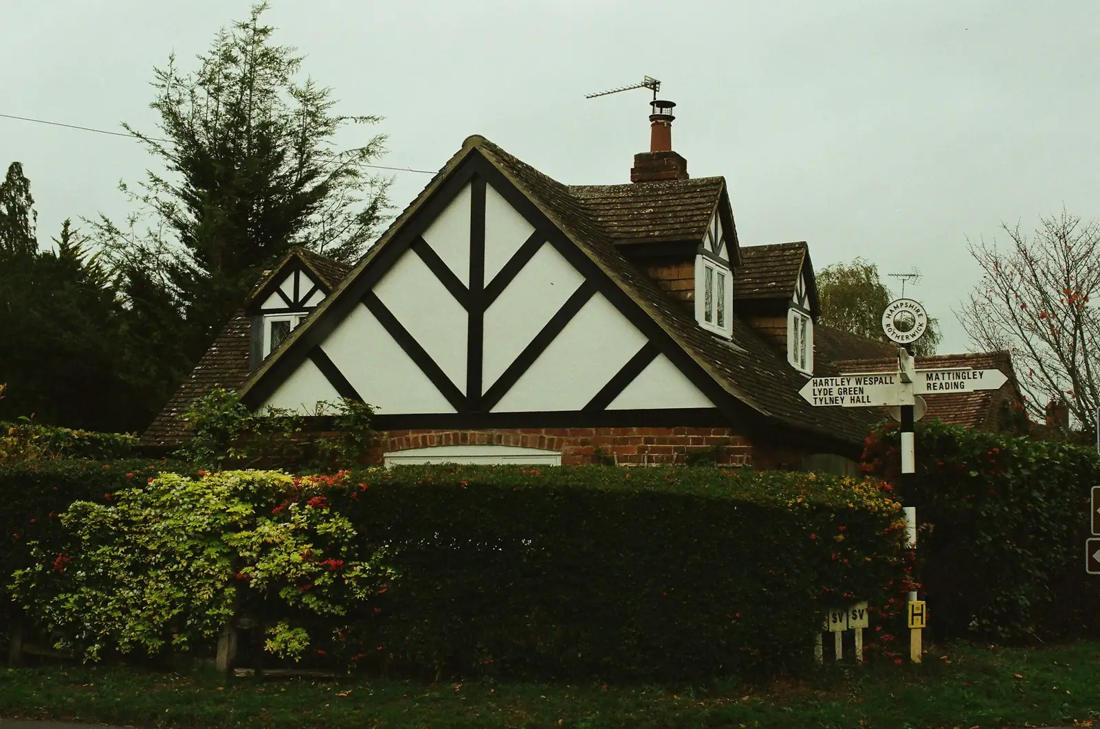 Black timber framing patterns the white gable end above brickwork, with a fingerpost at the hedge.