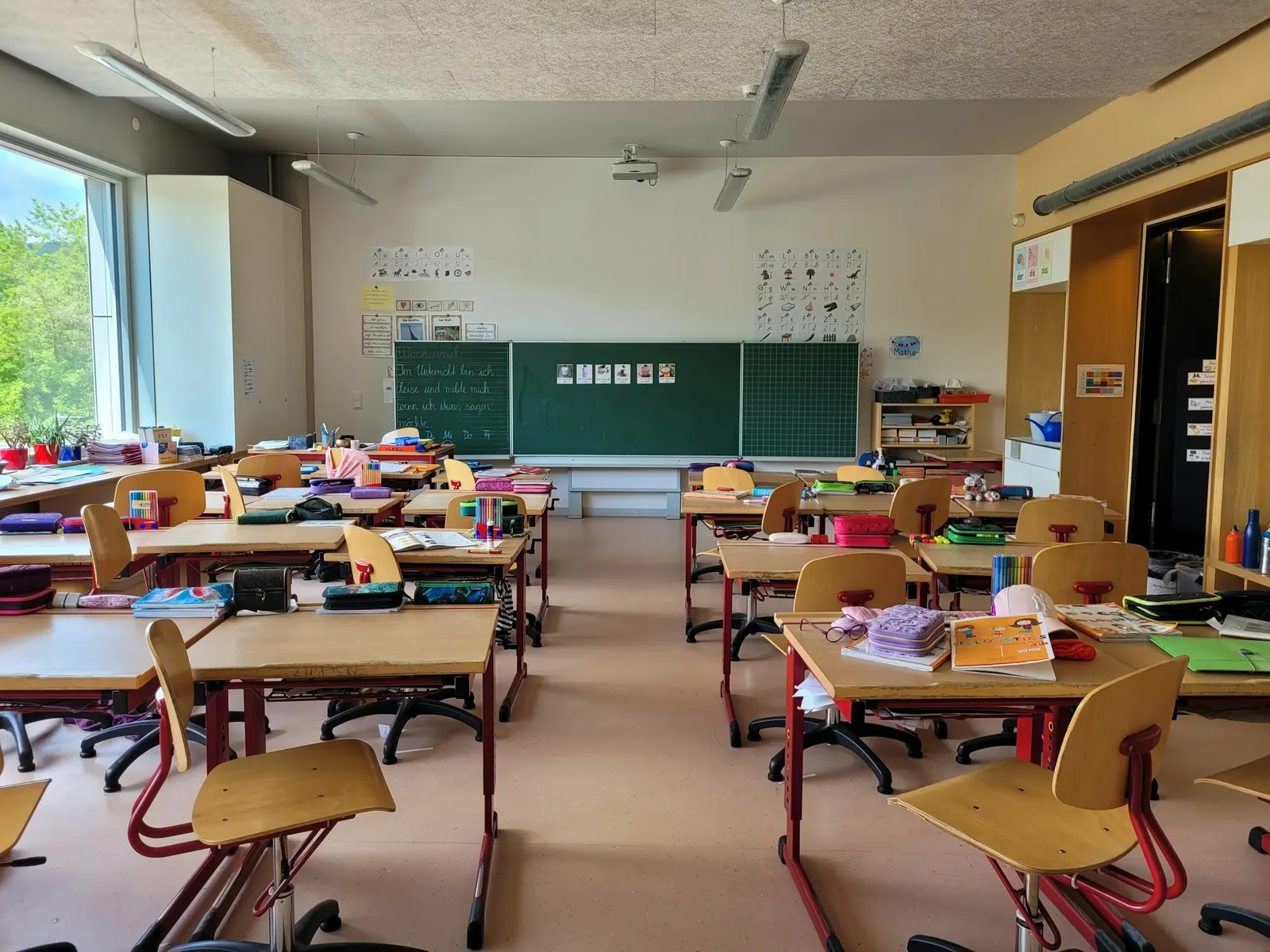 Height-adjustable desks on red steel frames face a green chalkboard, with strip lights and a projector above.