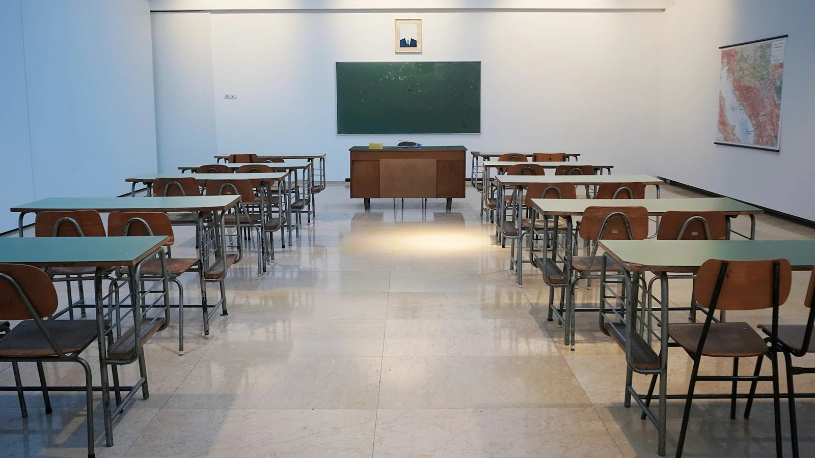 Green-topped tables and metal-framed chairs stand on polished stone, blackboard and teacher's desk at the front.