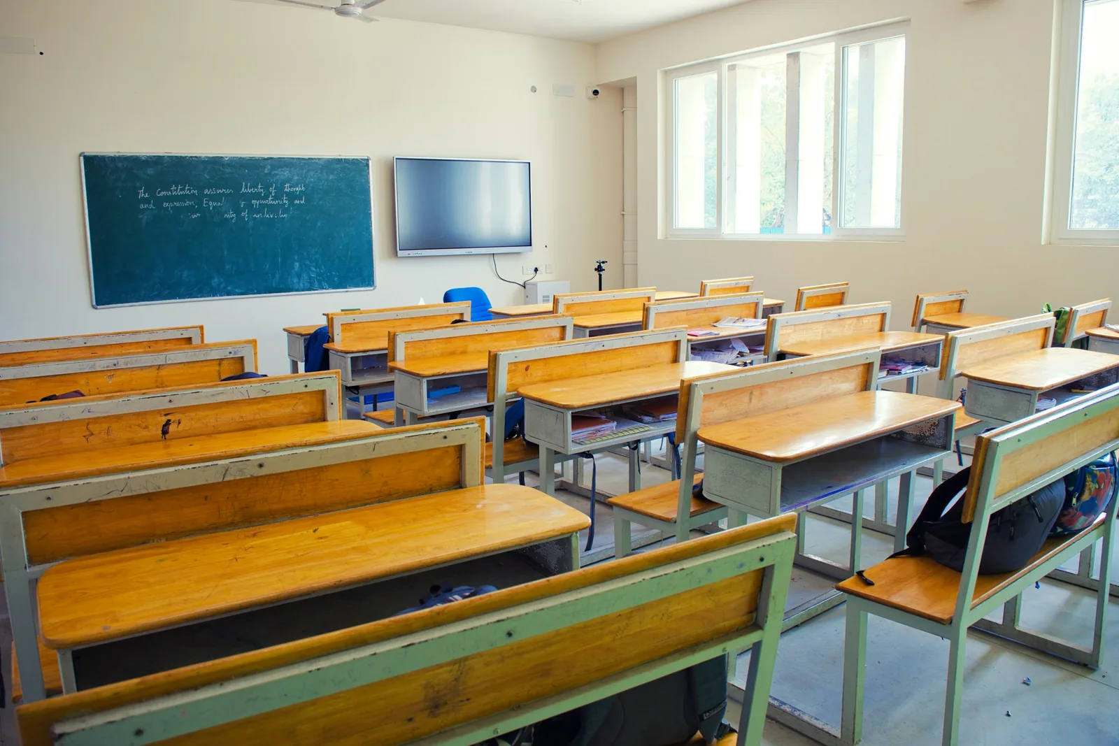 Timber bench desks sit on painted steel frames, bags dumped beneath, a screen mounted beside the board.