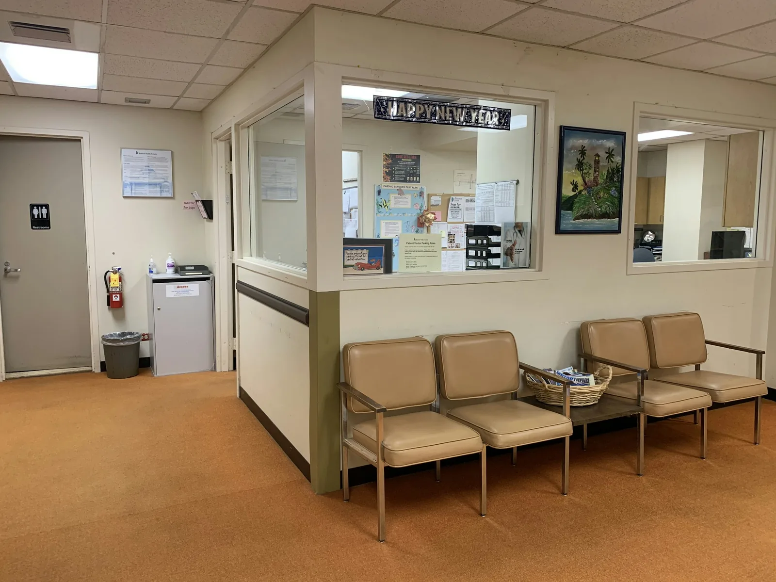 An older hospital reception: glazed hatch, tan vinyl chairs, orange sheet flooring, mineral-tile ceiling.