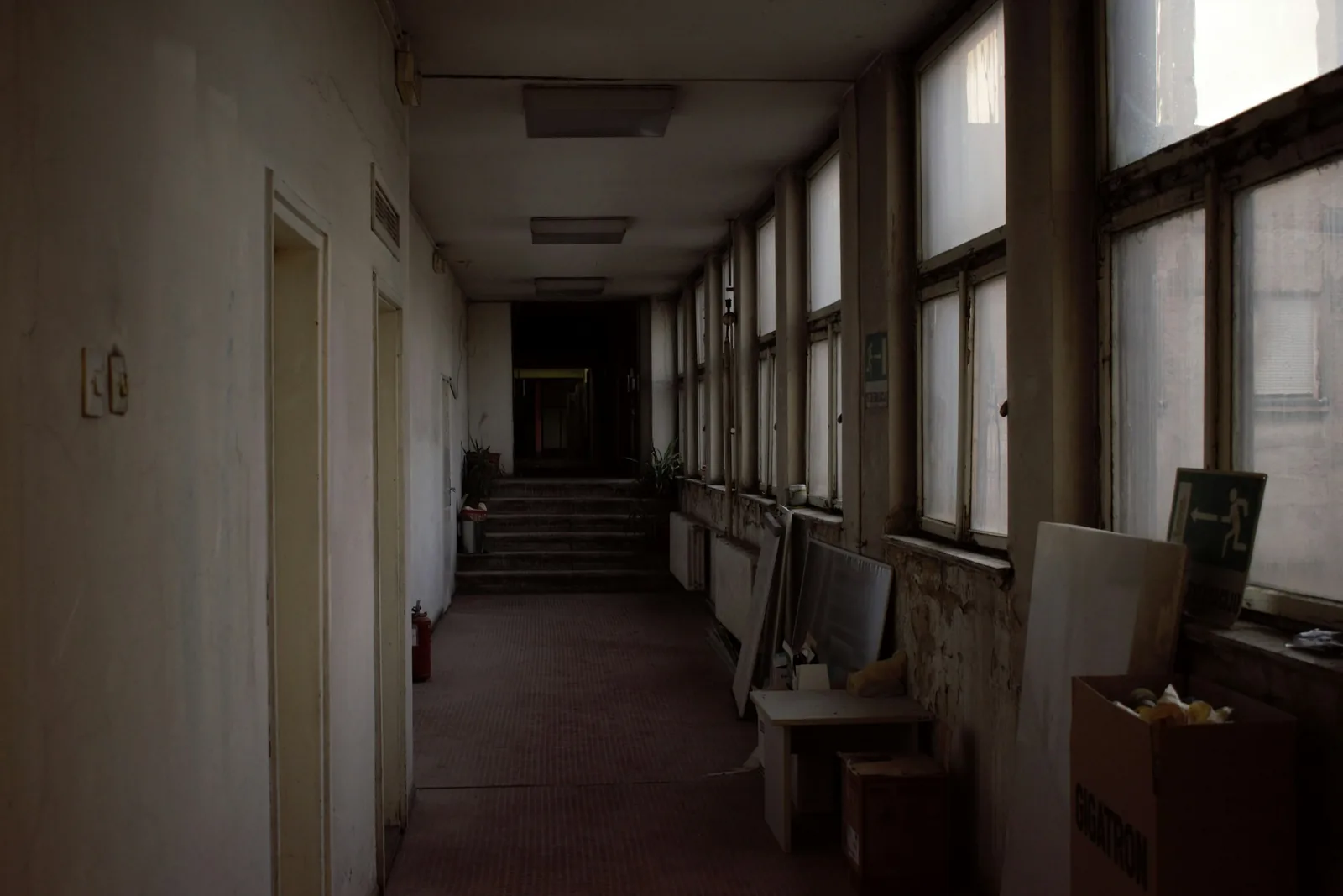 Boards and cardboard boxes stacked against the window wall of a dim corridor, radiators under the sills.