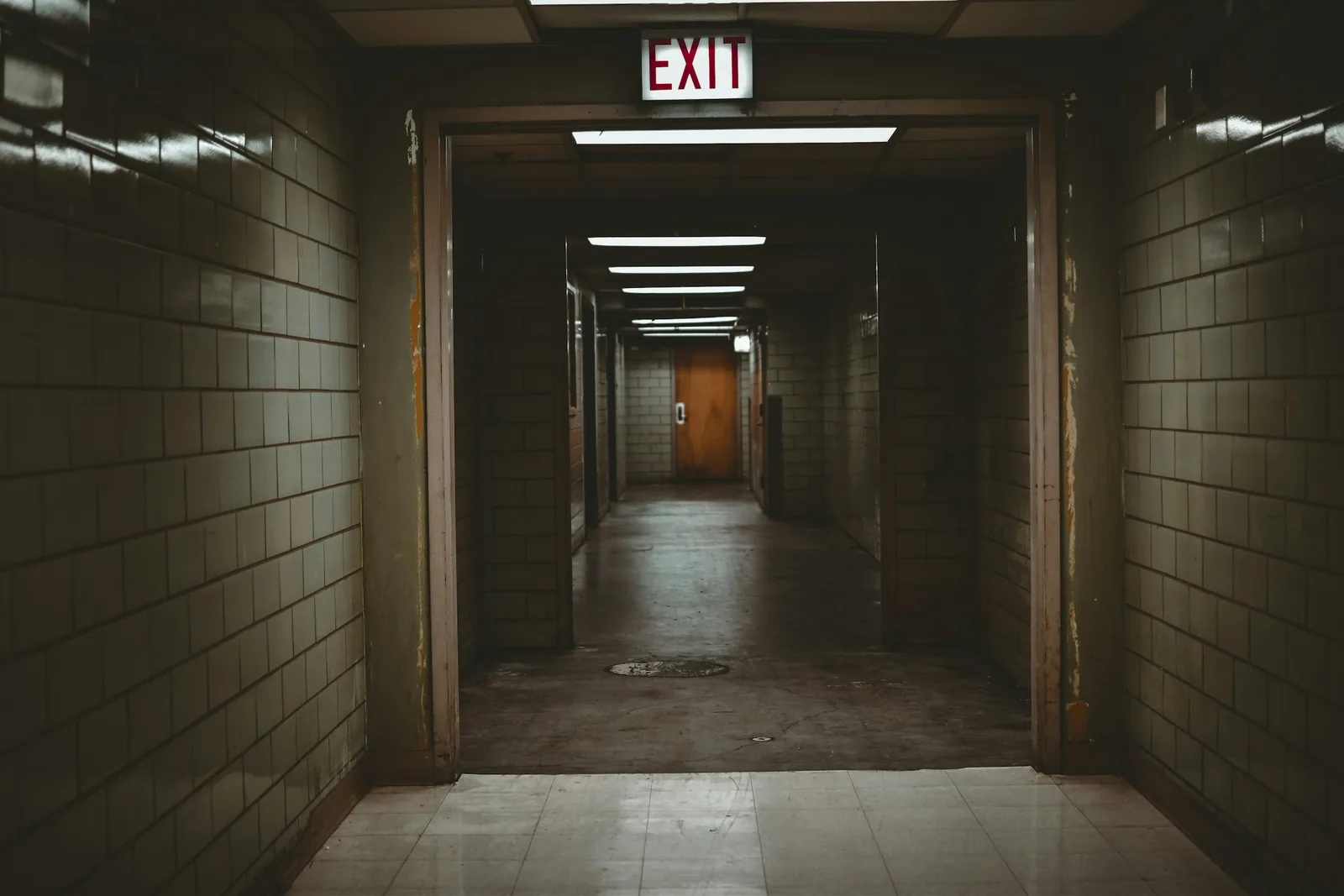 EXIT lettering glows over the opening into a glazed-brick corridor and a timber door beyond.