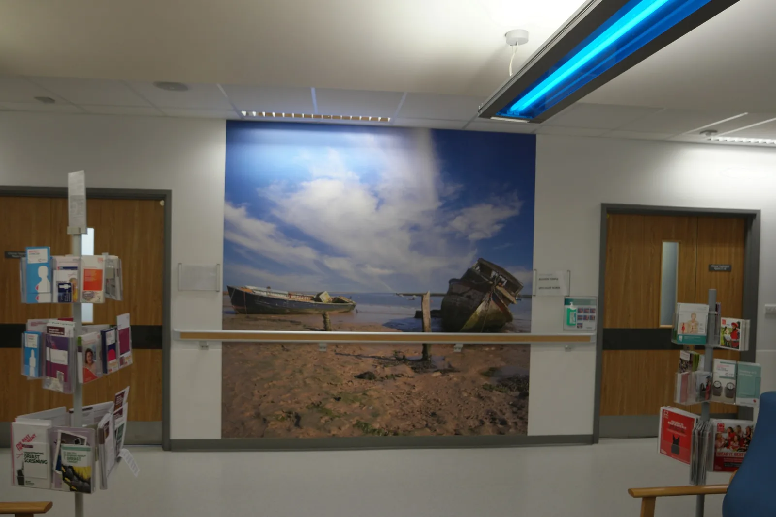 A finished community hospital waiting area with timber doors, a handrail and a large coastal photograph on the wall.