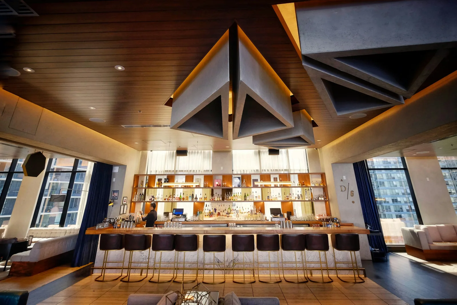 Faceted grey ceiling panels hang above a bar counter with a marble front and brass-framed stools.
