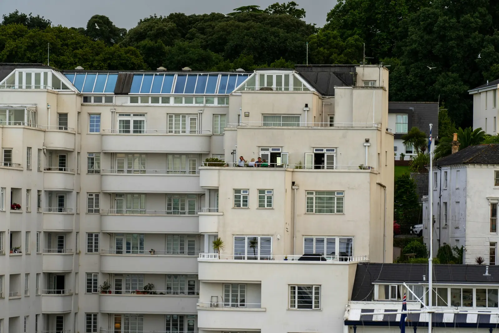 Woods rise behind a block of cream-rendered flats where four people sit out on a balcony.