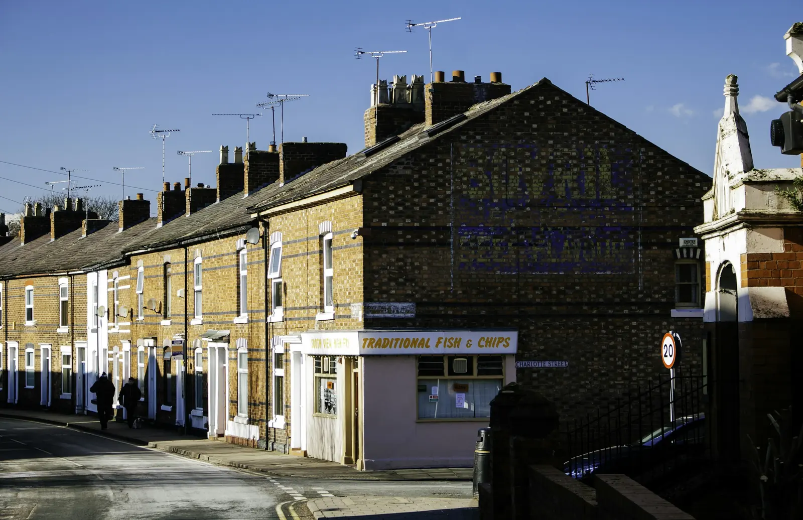 Aerials crowd the chimney stacks above a buff brick terrace that ends at a corner chip shop.
