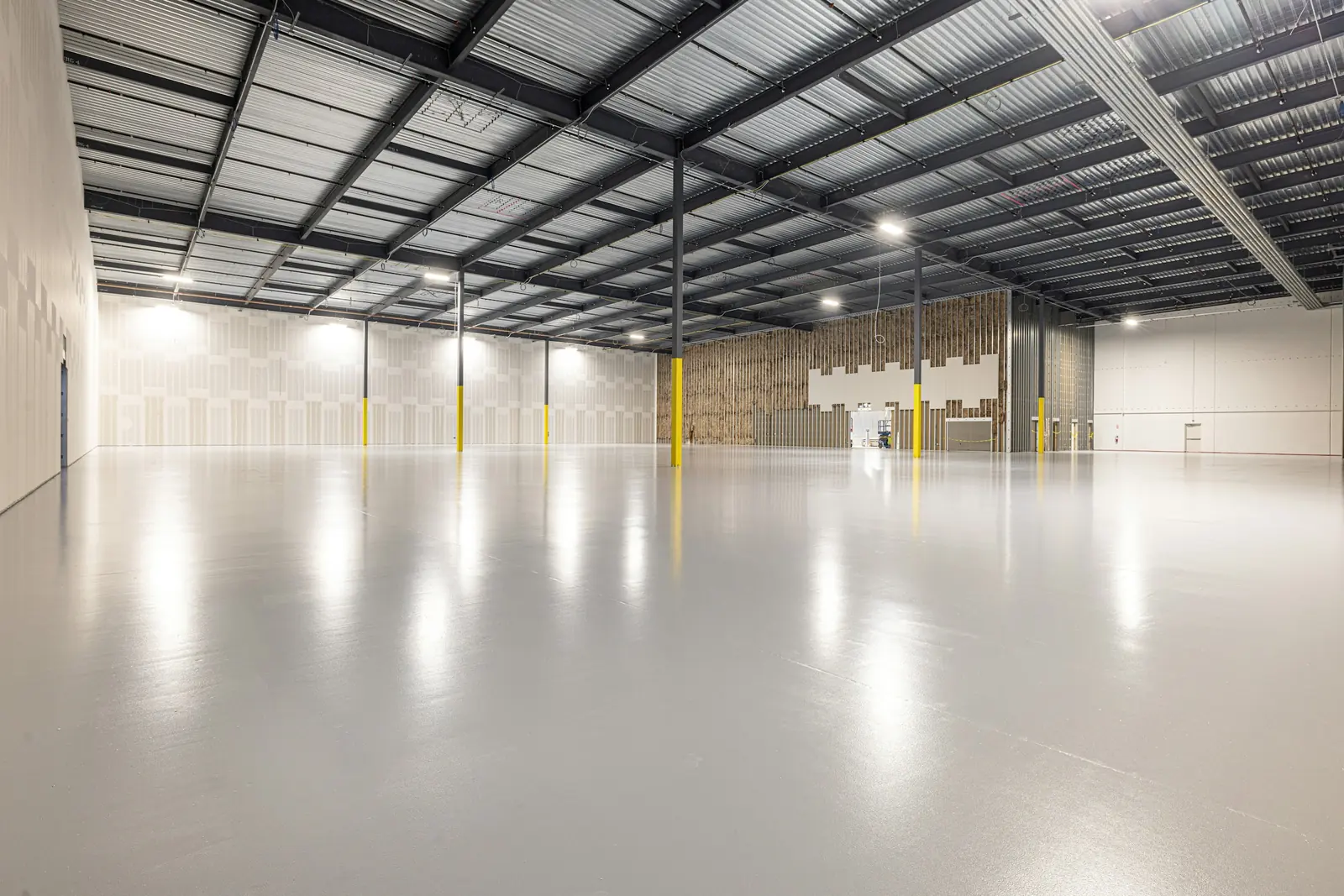 An empty warehouse with a freshly sealed pale grey floor, steel roof trusses and yellow-painted columns.