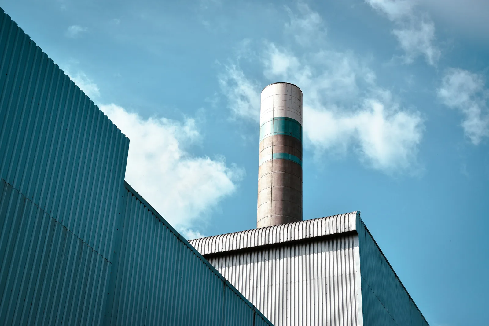 The profiled metal cladding of an industrial building, with a tall chimney stack against a blue sky.