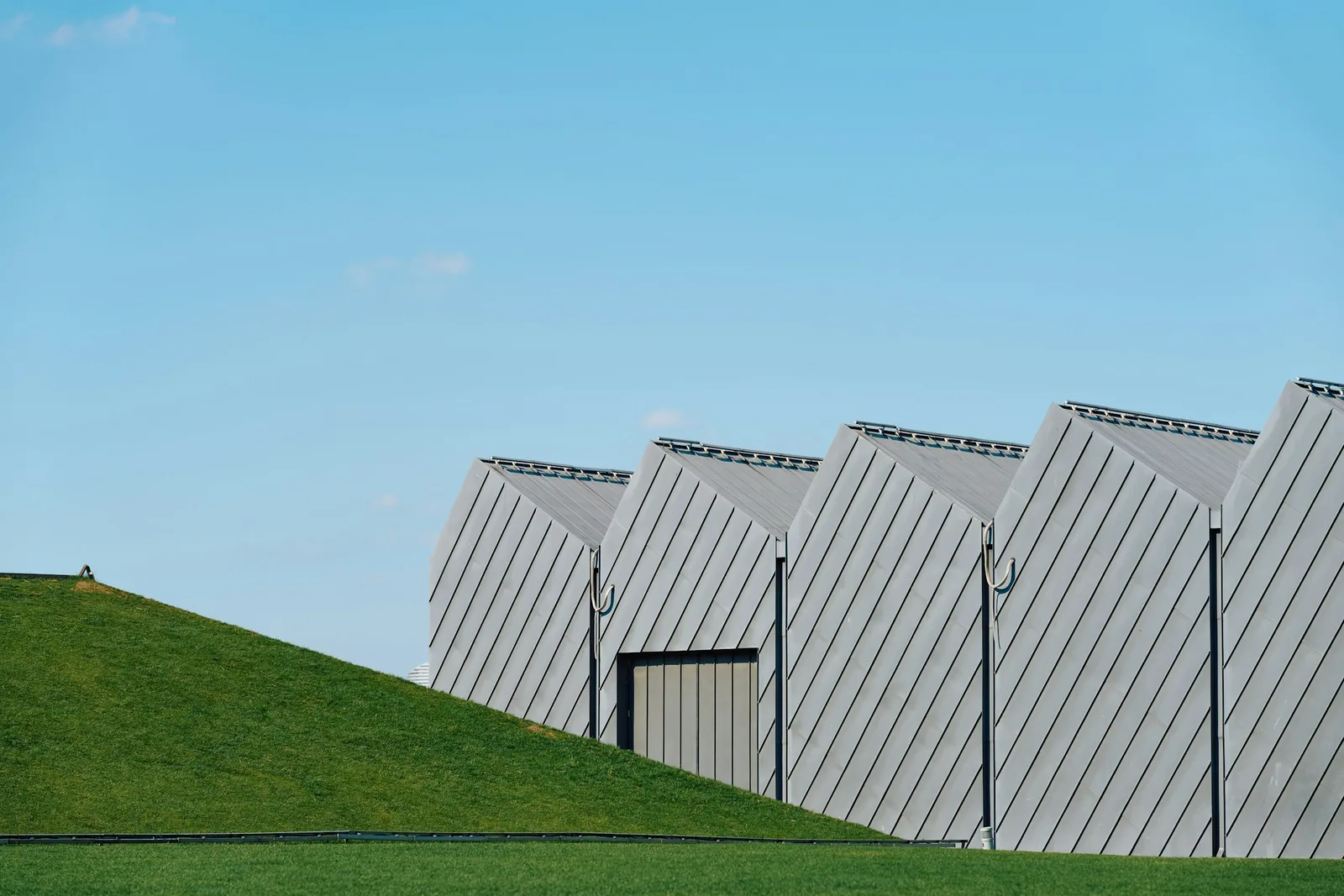 A modern industrial building in pale profiled cladding, with a sawtooth roofline and a roller shutter door, above a grass bank.