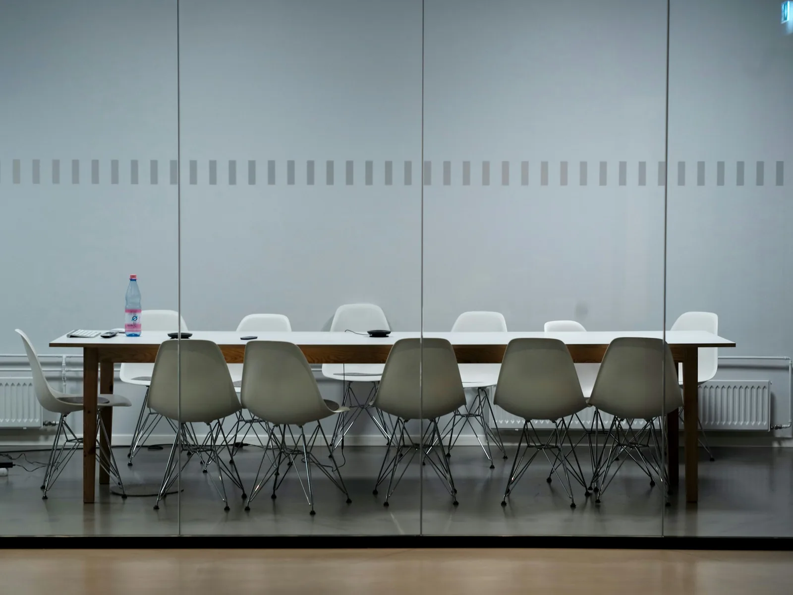 Behind a full-height glass partition, moulded plastic chairs on wire legs ring a long white table.