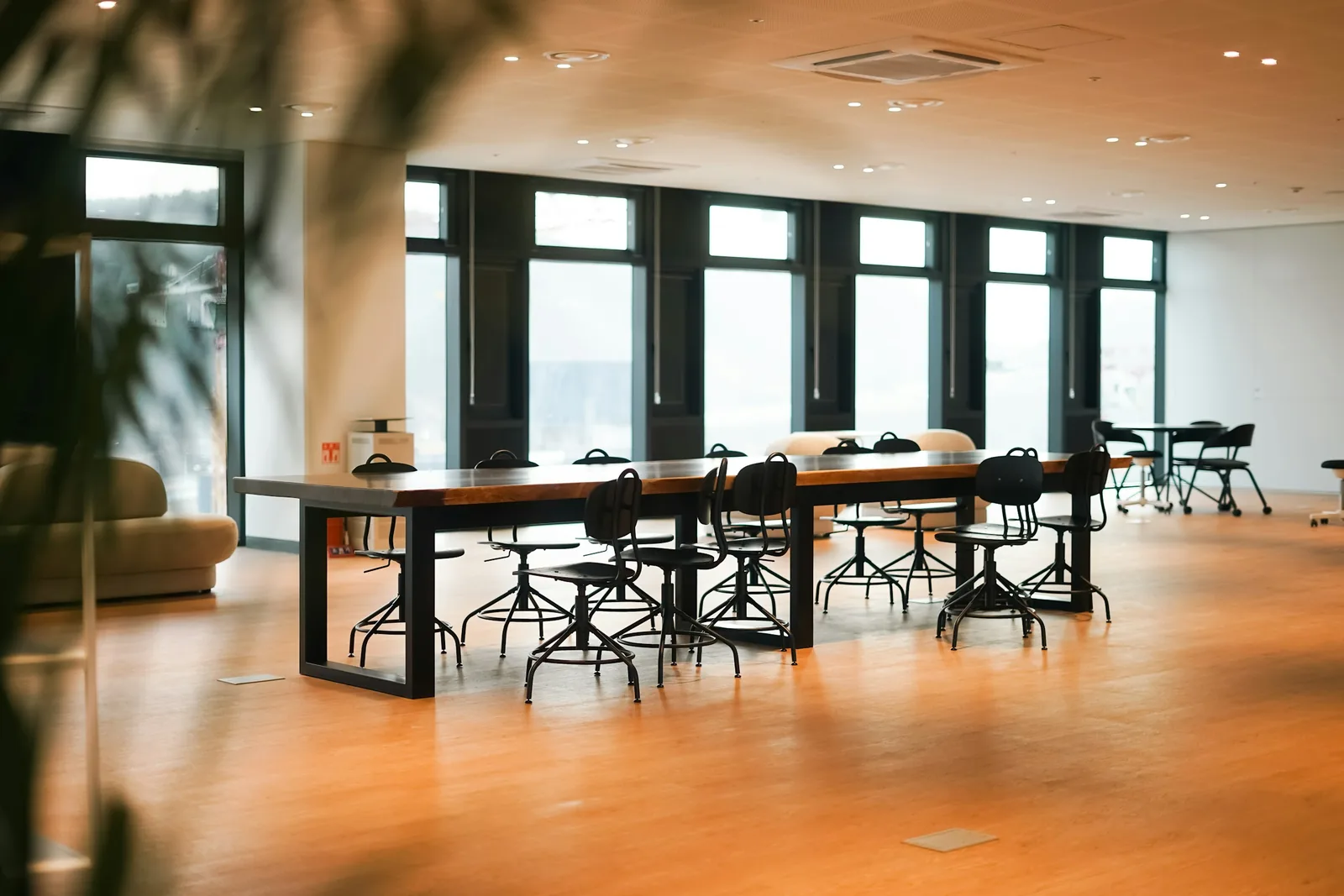 Black drafting stools pull up to a long timber-topped bench table on honey-coloured flooring.