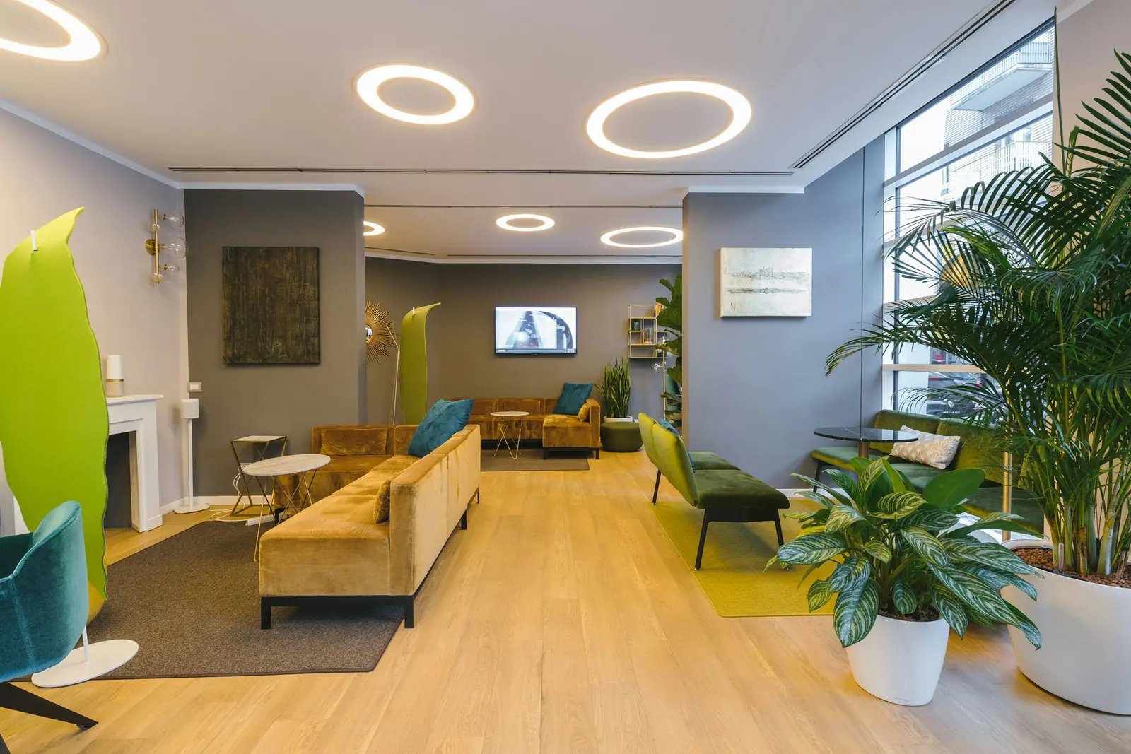 Ochre velvet sofas face a green bench across oak-look boards, ring lights set into the ceiling.