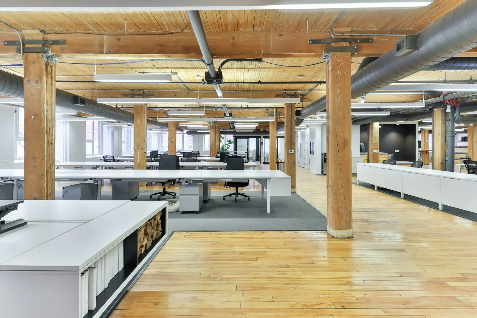 Exposed ductwork threads between heavy timber posts above white bench desks and a grey carpet island.