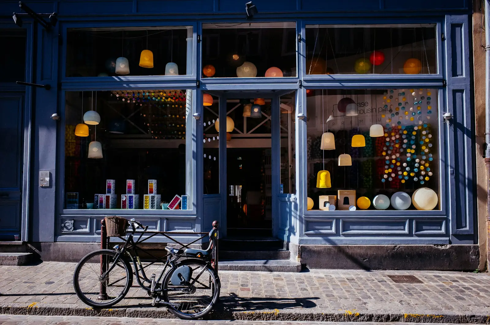 A bicycle leans at the kerb outside a blue timber shopfront hung with woven shades and coloured balls.