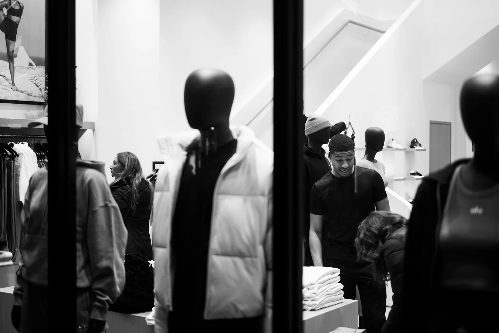 Black and white, shot through the glazing: a mannequin in a padded gilet, shoppers, trainers on wall shelves.