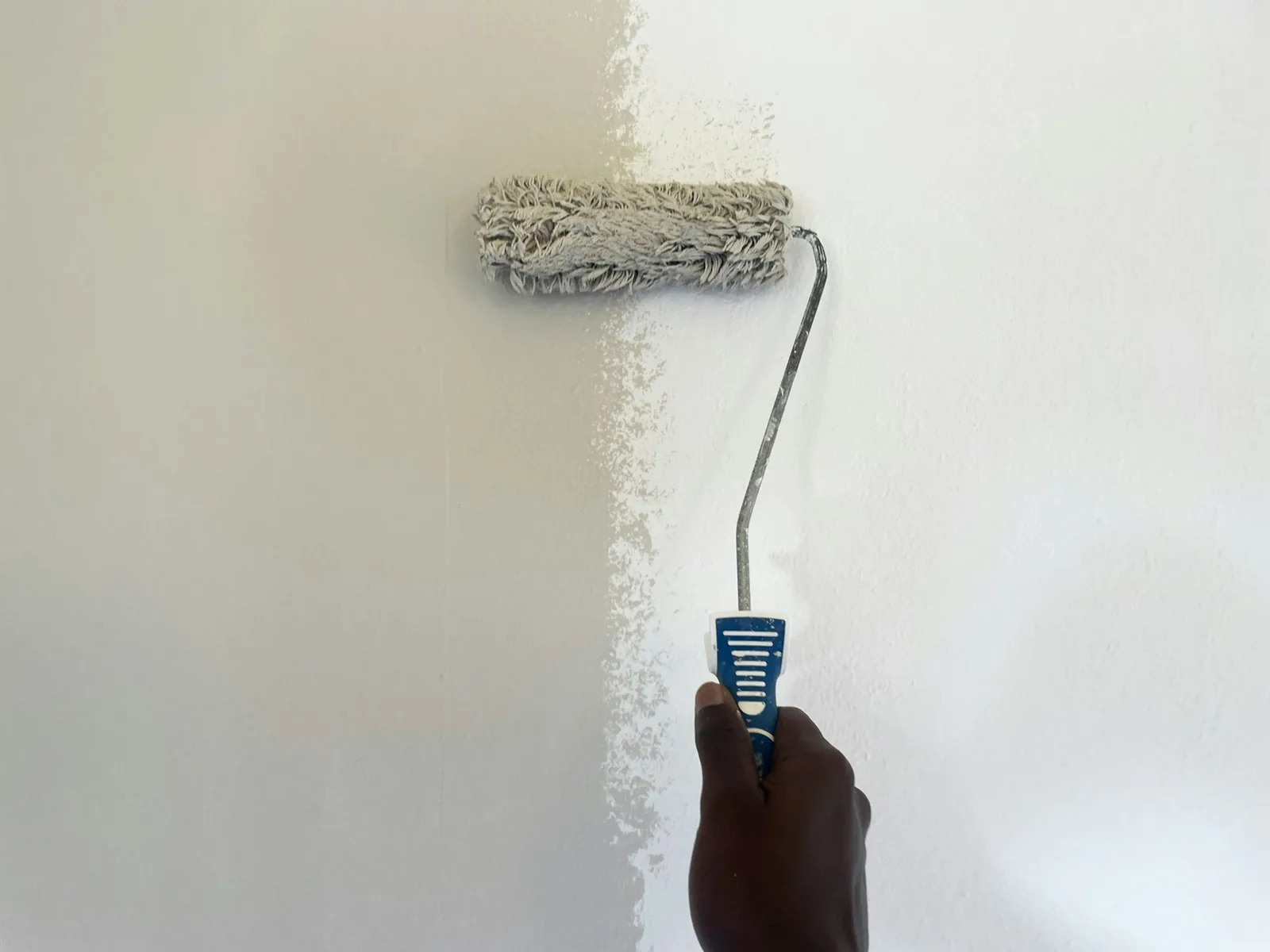 A hand paints a white wall; the roller is leaving a thin patchy stripe, not solid cover.