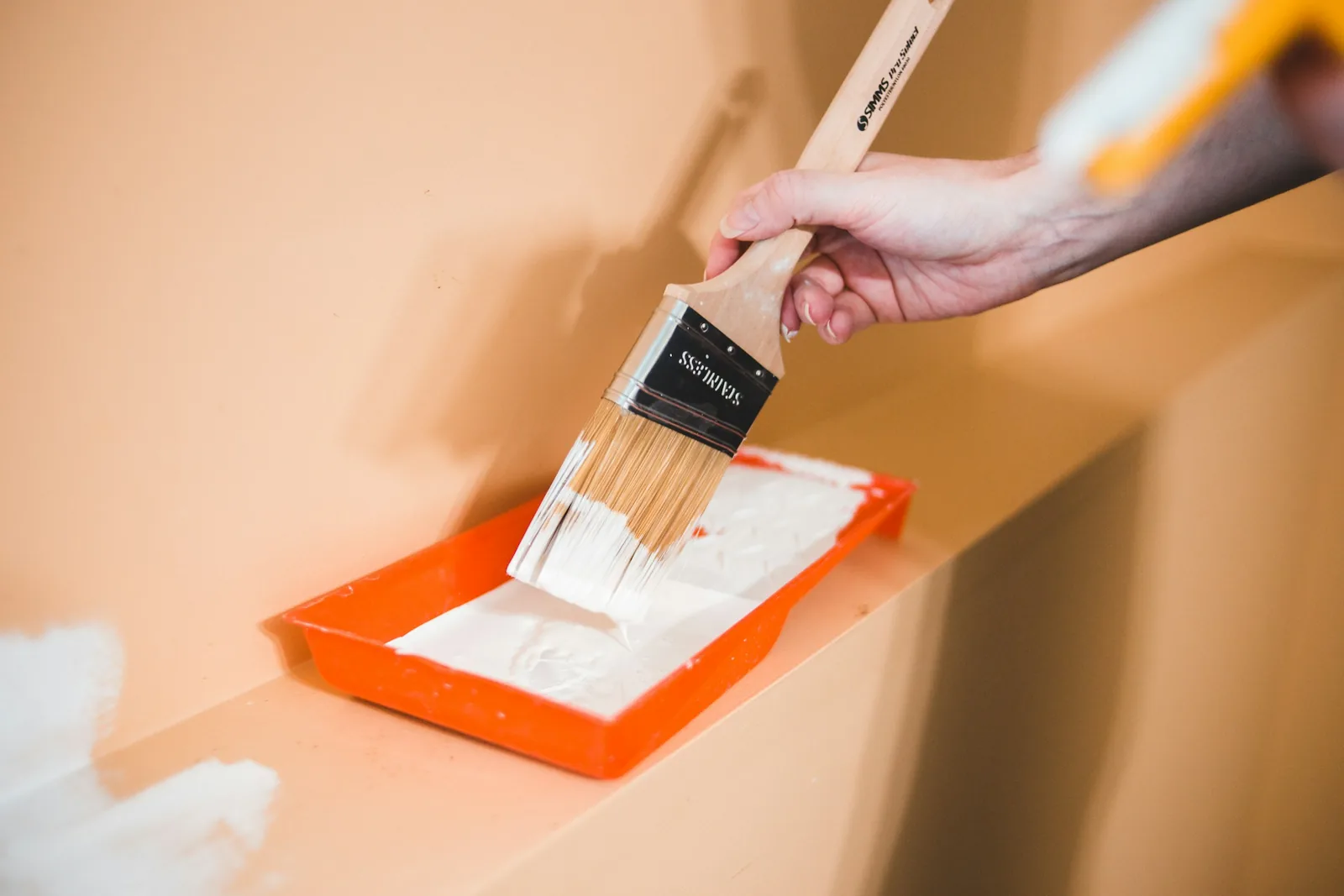 A hand loading a paintbrush from a paint tray beside a part-painted wall.